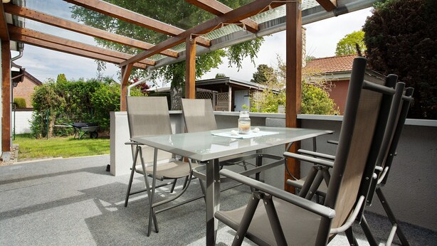 Une terrasse extérieure avec une table en verre et des chaises en métal, entourée d’une structure de pergola en bois.