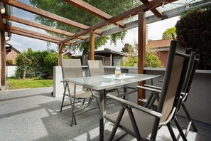 Une terrasse extérieure avec une table en verre et des chaises en métal, entourée d’une structure de pergola en bois.