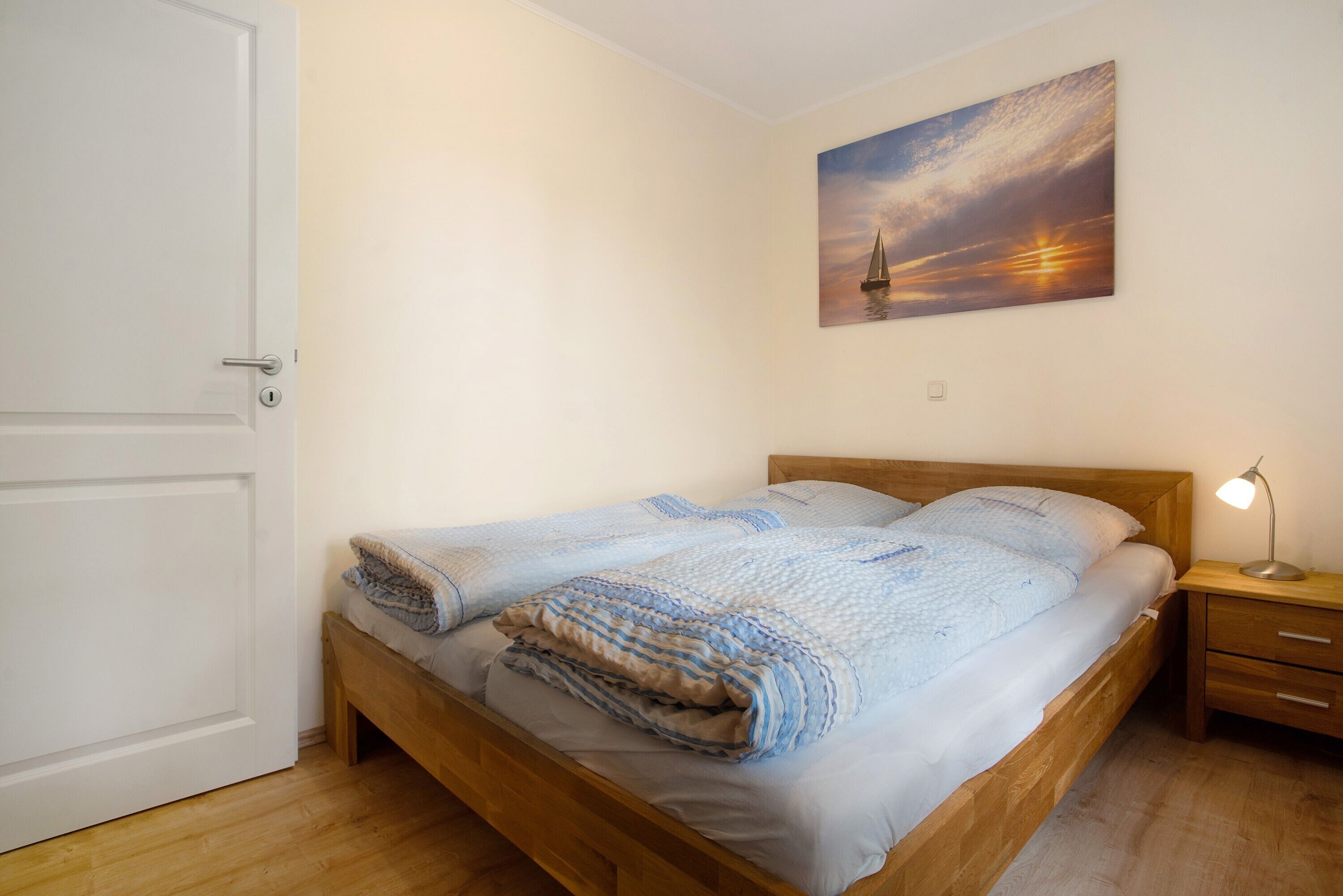 Un lit simple avec un cadre en bois, une table de chevet avec une lampe et un tableau représentant un coucher de soleil accroché au mur.