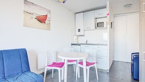 Un espace de vie compact comprenant un canapé bleu, une table à manger blanche avec quatre chaises, un coin cuisine avec des meubles blancs et un micro-ondes, ainsi qu’un tableau sur le thème de la plage accroché au mur.