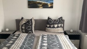 A bedroom with a checkered bedspread, two heart-patterned pillows, a painting of a coastal scene, and a view of the sea. - Apartment 'Orthsee 2' with Shared Terrace and Wi-Fi (Hohen Wangelin)