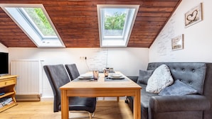 Een eethoek met een houten tafel, stoelen en een grijze bank. Boven de tafel zijn dakramen en aan de muur staat een radiator.