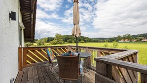 Een houten terras met een tafel en stoelen, een hangende parasol, en een uitzicht op een groen veld en huizen in de verte.