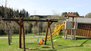 A playground with swings and a slide, surrounded by trees and a building.