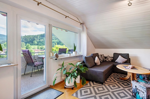 Une pièce chaleureuse avec un canapé gris, une petite table ronde, une plante en pot et une vue sur un paysage verdoyant par une grande fenêtre.