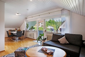 Un intérieur moderne comprenant un coin repas, un canapé et de grandes fenêtres donnant sur un paysage verdoyant et des bâtiments.