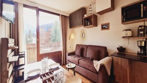 Ein gemütliches Wohnzimmer mit einem braunen Sofa, einem Esstisch mit Stühlen, einer Kaffeemaschine und einem Fernseher an der Wand.