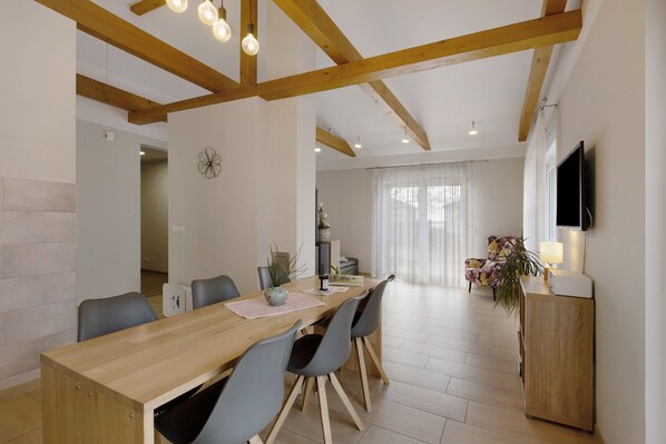 A dining area with a wooden table, grey chairs, a TV mounted on the wall, and a plant on a cabinet.