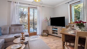 A living room with a sofa, a TV, a wooden dining table, and a window with a view of trees. - Apartment 'Spreequartier' with Lake View, private Terrace and Wi-Fi (Malschwitz)