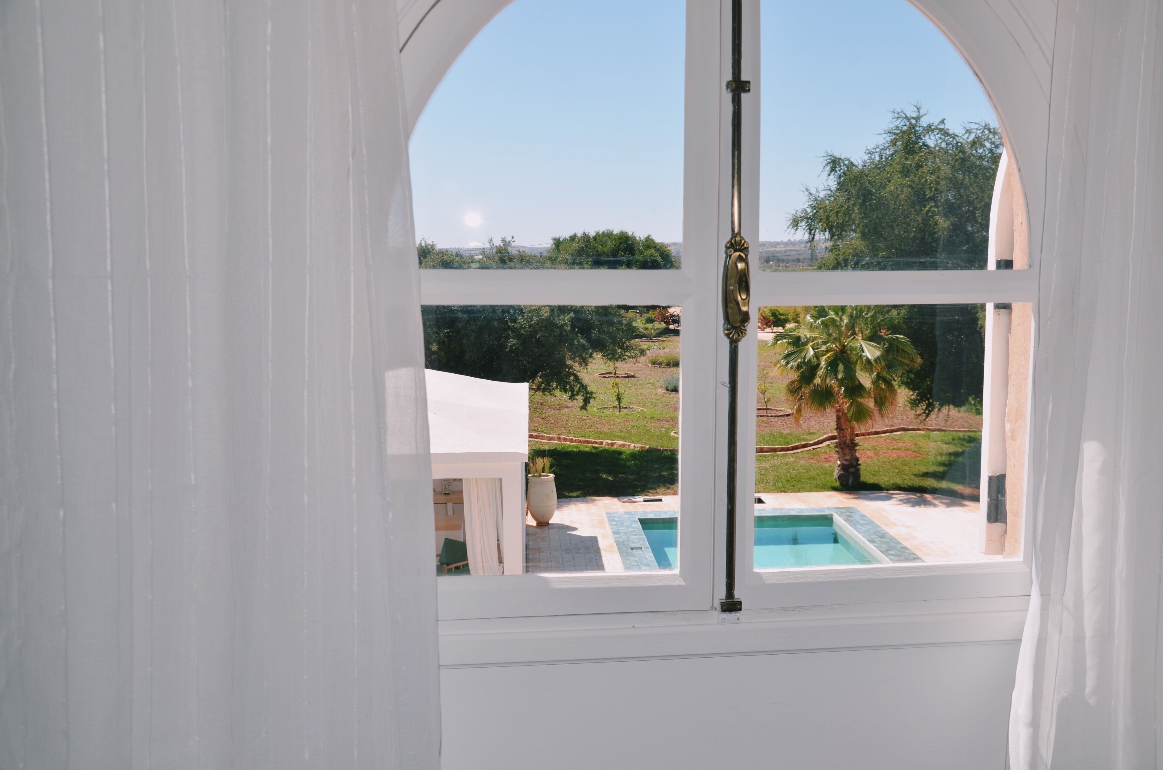 Vista da una stanza bianca con un'ampia finestra che si affaccia su una piscina e un giardino curato.