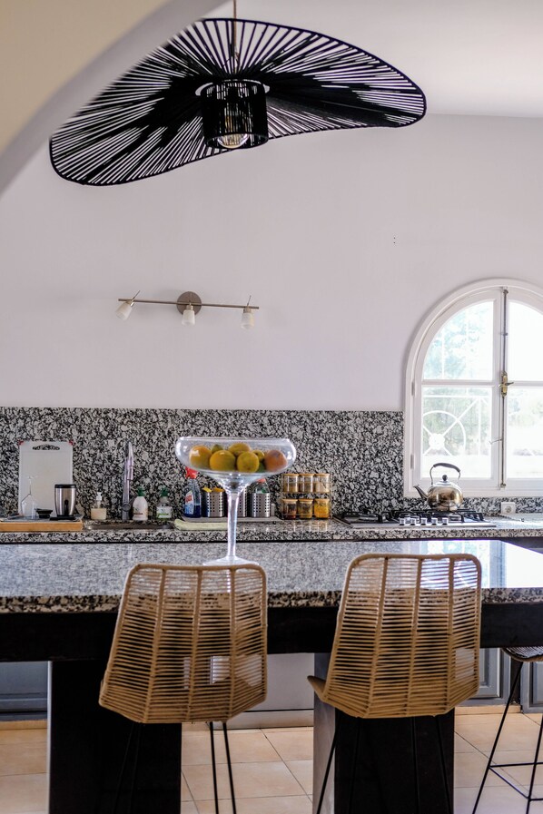 A kitchen with a large island, two wicker chairs, a hanging light fixture, and a window with a view. - A haven of peace in the countryside surrounded by 90 olive and 10 argan trees (Essaouira)