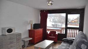 Una sala de estar con un sofá, una butaca roja, una mesa de centro de madera, un televisor sobre un mueble, un armario rojo y un microondas.