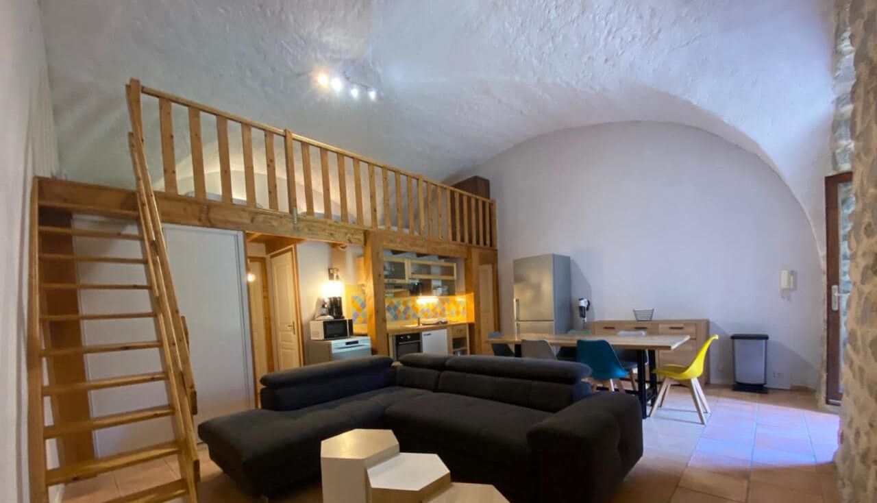Un espace de vie chaleureux avec une mezzanine, un canapé, une table à manger et une cuisine en arrière-plan.