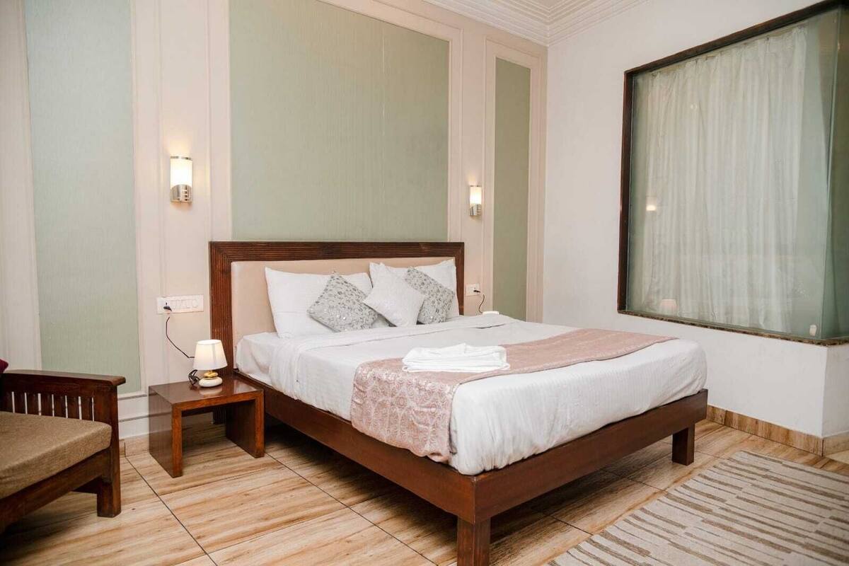 A hotel room with a wooden bed, a nightstand with a lamp, a chair, and a large window with curtains.