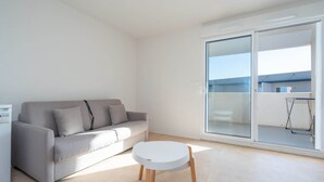 Ein modernes Wohnzimmer mit einem grauen Sofa, einem kleinen weißen Tisch und einer Glasschiebetür, die auf einen Balkon führt.