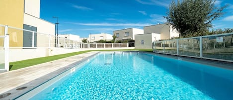 A rectangular swimming pool with clear blue water, surrounded by a white fence and a grassy area.