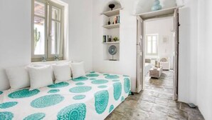 A bedroom with a bed, pillows, a bedside table, a shelf with books, and a door leading to another room.
