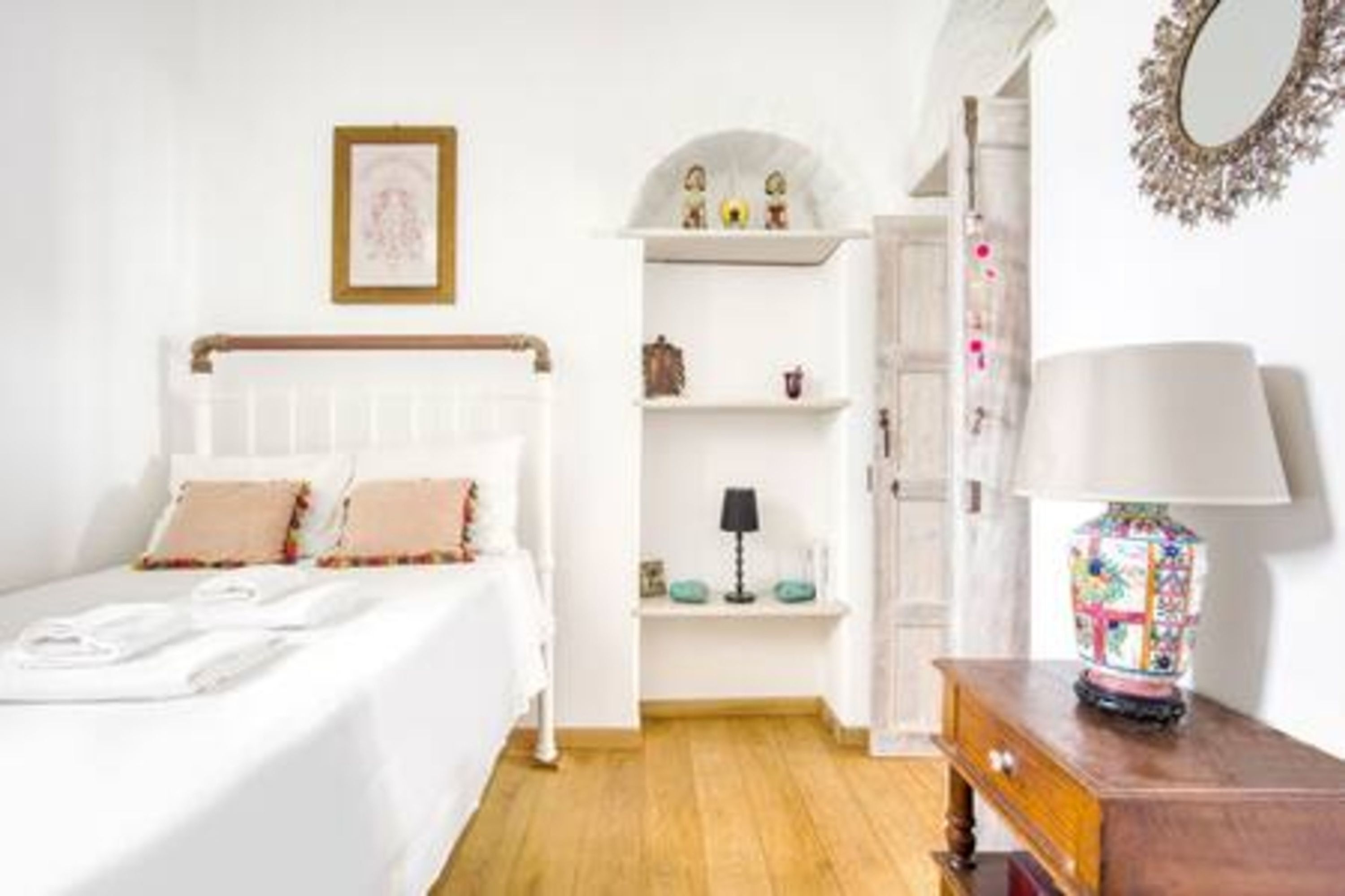 A bedroom with a bed, a nightstand, a dresser, a shelf with decorative items, and a mirror on the wall.