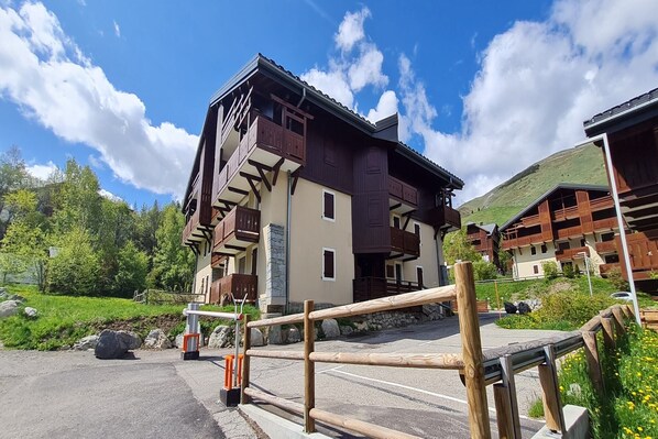 Un immeuble à plusieurs étages, doté de balcons, d’un rez-de-chaussée en pierre et d’un espace de stationnement délimité par une clôture en bois.