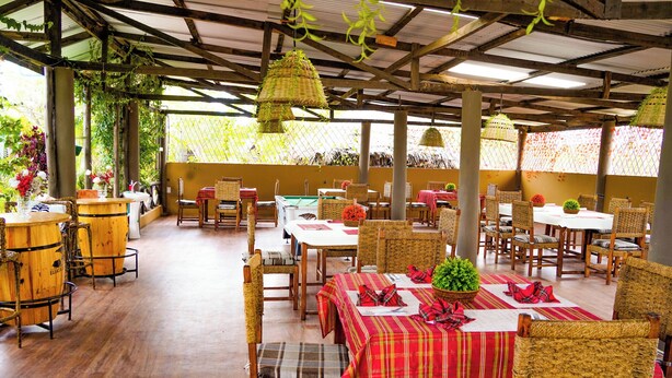 An outdoor dining area with wicker chairs and tables, covered by a metal roof, and surrounded by greenery. - Eliata Bush Camp (Kajiado)