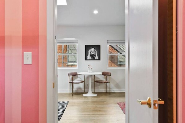 Una habitación con una puerta blanca, una mesa redonda con dos sillas, un cuadro blanco y negro en la pared y una ventana con vistas a un edificio de ladrillo.
