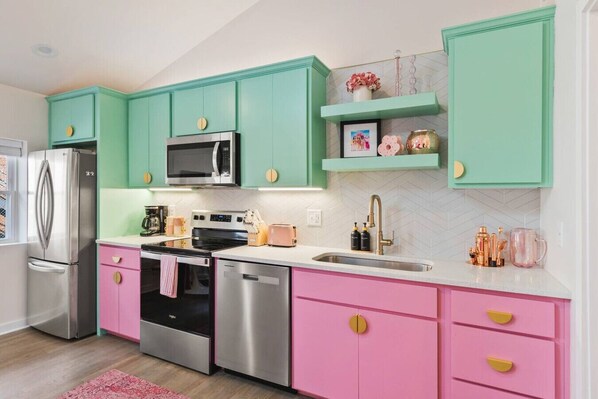 Una cocina con armarios azul verdoso, cajones rosados, electrodomésticos de acero inoxidable y encimera blanca.