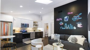 A modern living room with a grey sofa, white armchairs, and a coffee table. There is a neon sign on the wall and a kitchen in the background. - Vibrant Neon Condo Near Restaurants, Coffee & Bars (Nashville)