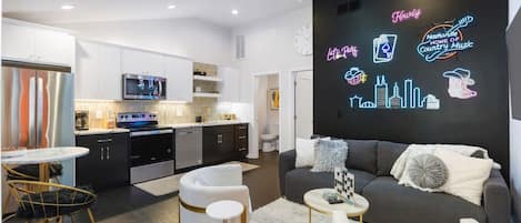 Una sala de estar moderna con un sofá gris, sillones blancos y una mesa de centro. Hay un letrero de neón en la pared y una cocina al fondo.