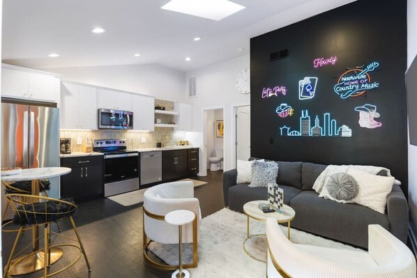 Una sala de estar moderna con un sofá gris, sillones blancos y una mesa de centro. Hay un letrero de neón en la pared y una cocina al fondo.