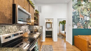Una cocina moderna con estufa de acero inoxidable, microondas y armarios de madera. Se ve un pasillo que conduce a un dormitorio con cama y pared verde con un mural floral.