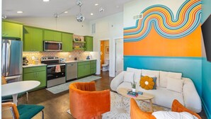 Una cocina moderna con gabinetes verdes y electrodomésticos de acero inoxidable, una zona de comedor con sillas naranjas y un salón con un sofá blanco y un mural colorido.