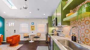 Una cocina moderna con gabinetes verdes, encimera de mármol y horno integrado. Hay un comedor con sillas anaranjadas y una mesa pequeña.