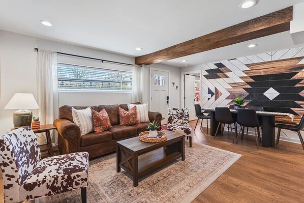A modern living room with a brown leather sofa, patterned armchairs, a wooden coffee table, and a dining area with a black and white geometric wall design.