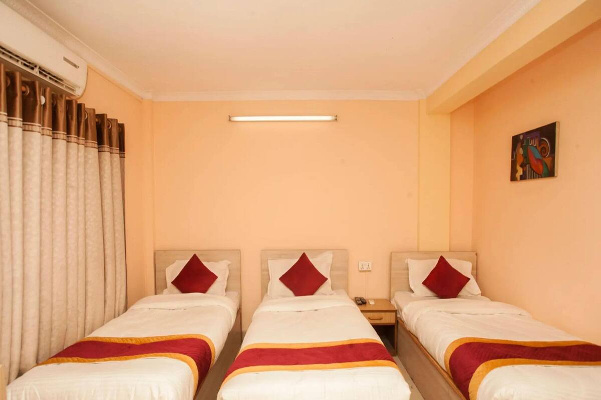 A hotel room with three beds, each with white bedding and red accents, a ceiling fan, and a painting on the wall.
