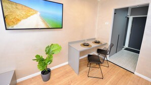 Una habitación moderna con televisión de pantalla plana mostrando una escena de playa, una pequeña mesa de comedor con sillas y una planta en maceta.