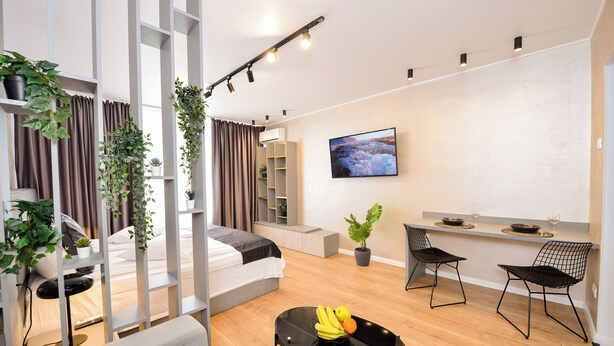 Una sala de estar moderna y bien iluminada con un sofá, una mesa de comedor con sillas, un televisor montado en la pared y plantas en macetas.