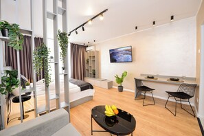 A modern, well-lit living room with a sofa, a dining table with chairs, a TV mounted on the wall, and potted plants.