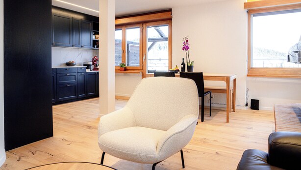 A modern living room with a sofa, a dining area with a table and chairs, and a kitchen with dark cabinets and a wooden countertop.