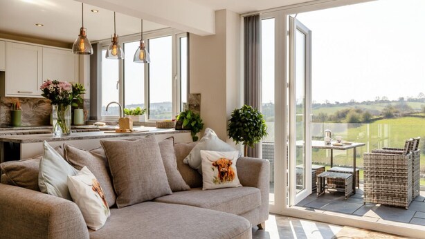 A modern living room with a sofa, a dining area with wicker furniture, and a kitchen with white cabinets and pendant lighting.
