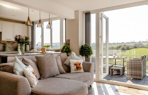 A modern living room with a sofa, a dining area with wicker furniture, and a kitchen with white cabinets and pendant lighting.