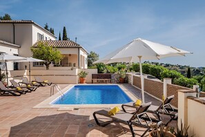 Ein Swimmingpool mit Liegestühlen und Sonnenschirmen, umgeben von einem Gebäude mit einem Ziegeldach und Blumentöpfen.