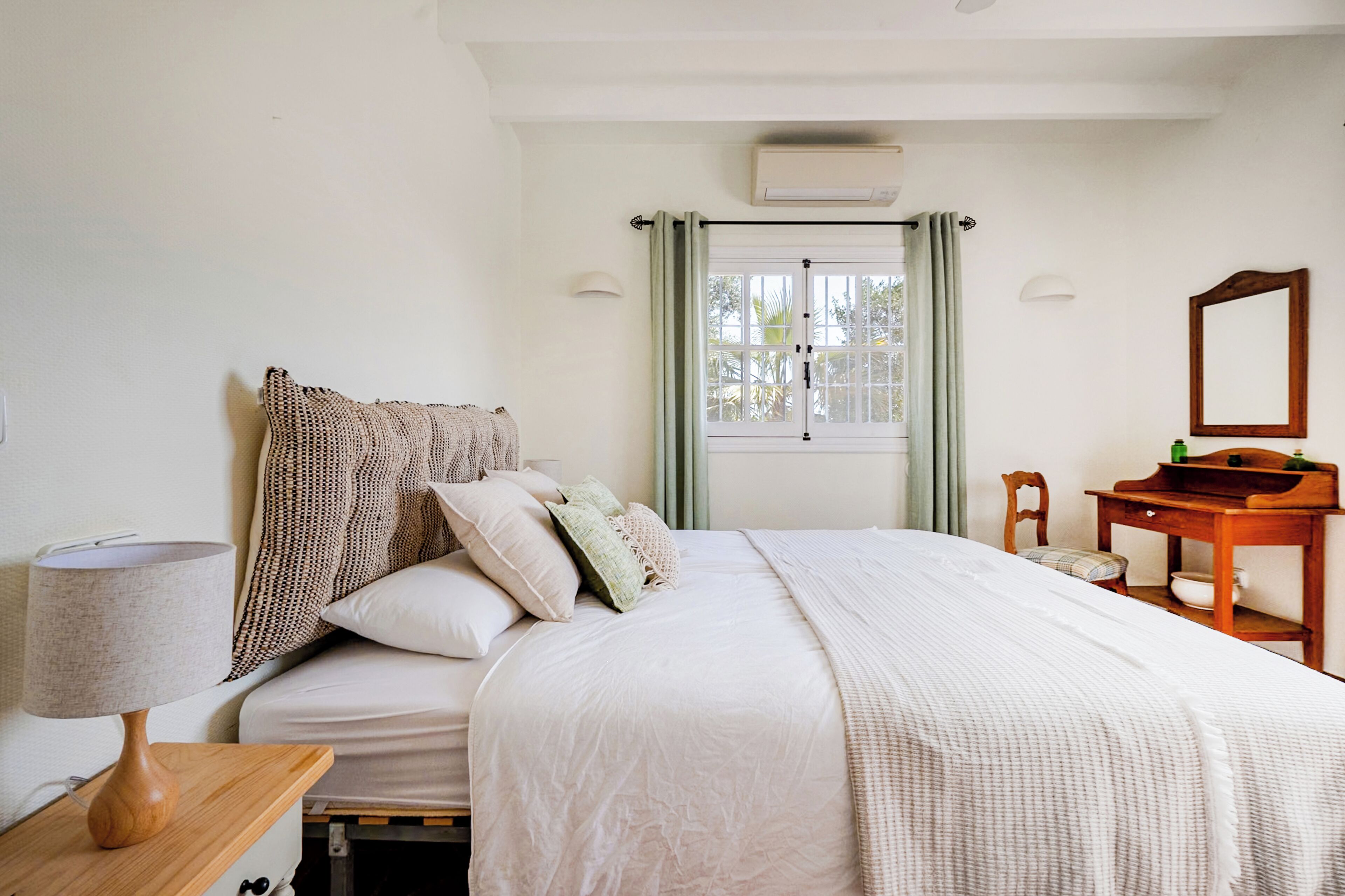 A bedroom with a bed, bedside table, lamp, window with curtains, and a mirror.