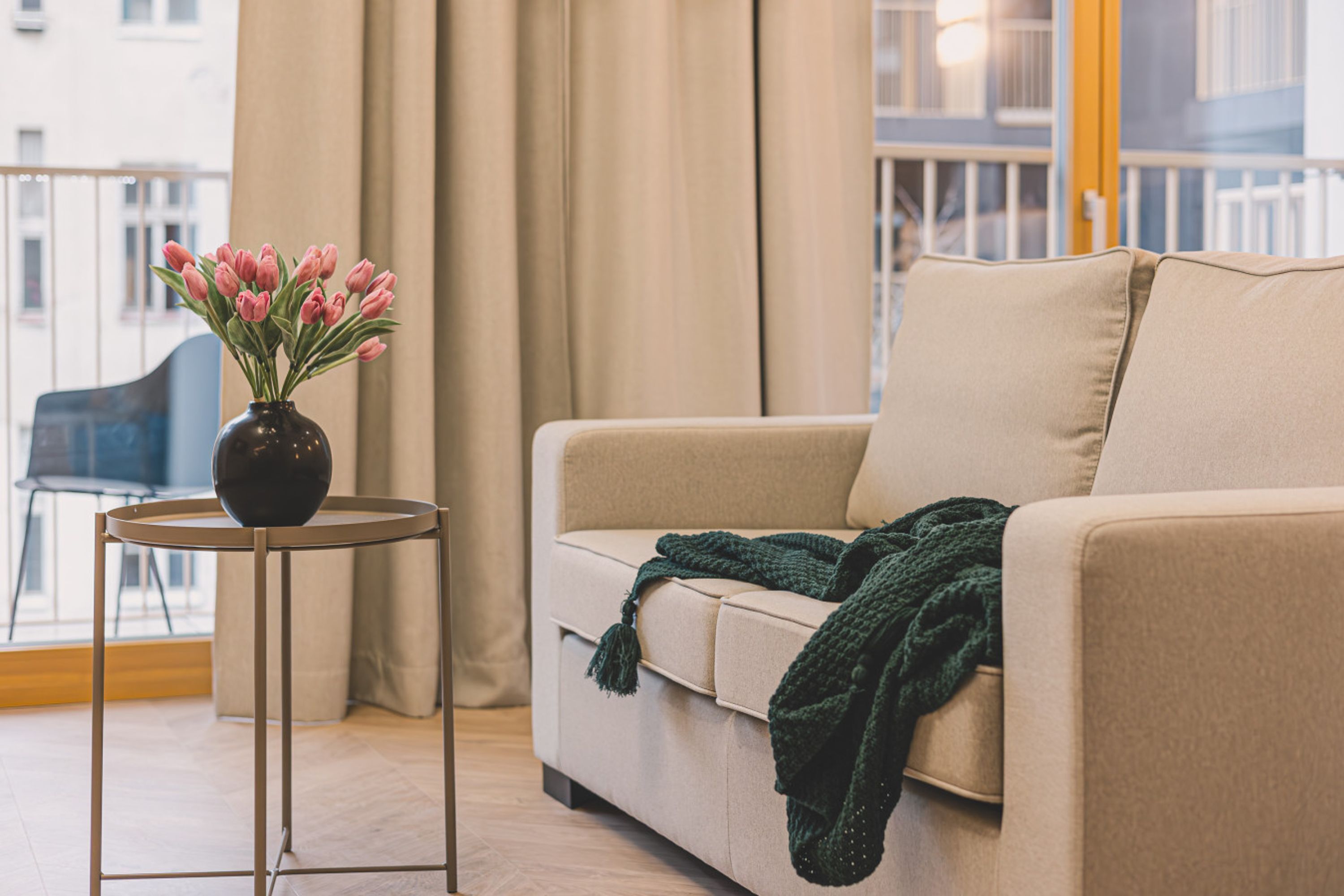 A living room with a beige sofa, a small round table with a vase of pink flowers, and a green throw blanket.