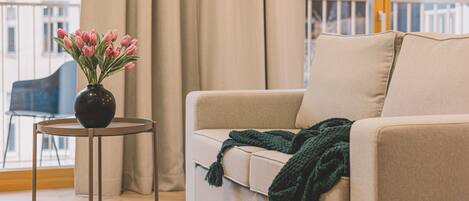 A living room with a beige sofa, a small round table with a vase of pink flowers, and a green throw blanket.