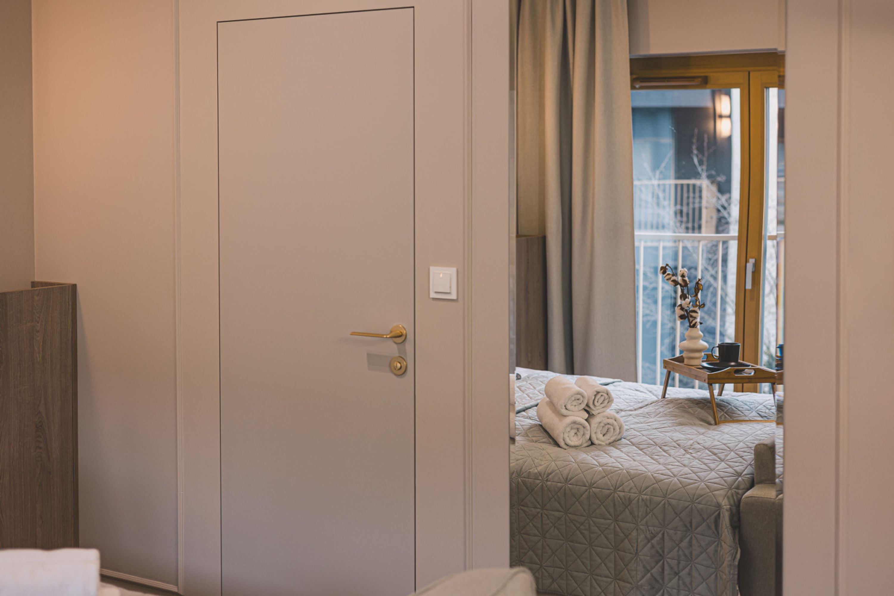 A hotel room with a closed door, a bed with rolled towels, a small table with a vase and a cup, and a window with curtains.