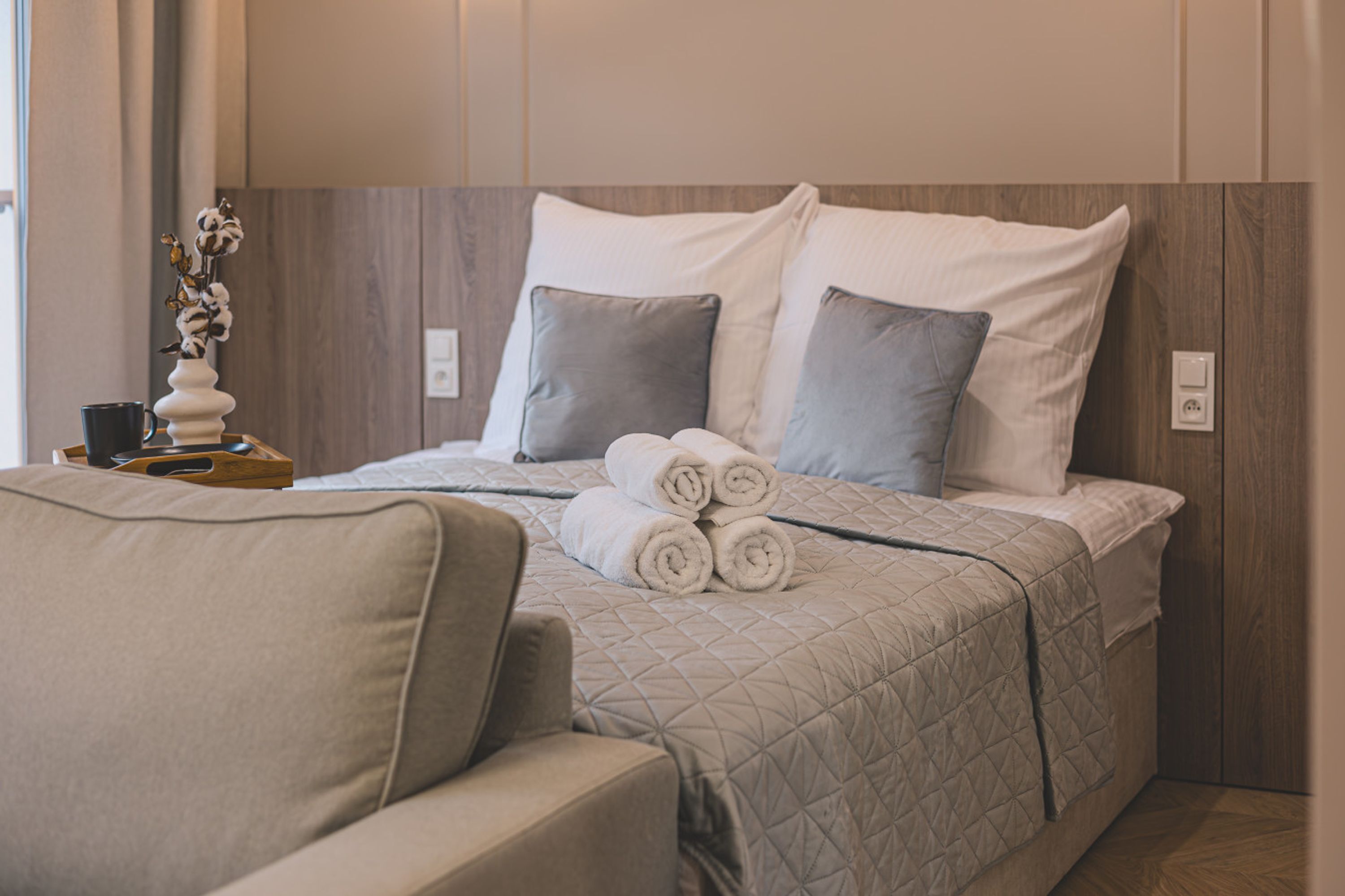 A neatly made bed with rolled towels, a bedside table with a vase, and a chair in a modern room with wooden paneling.