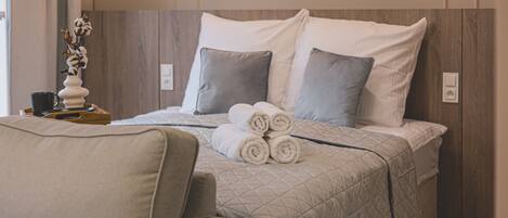 A neatly made bed with rolled towels, a bedside table with a vase, and a chair in a modern room with wooden paneling.