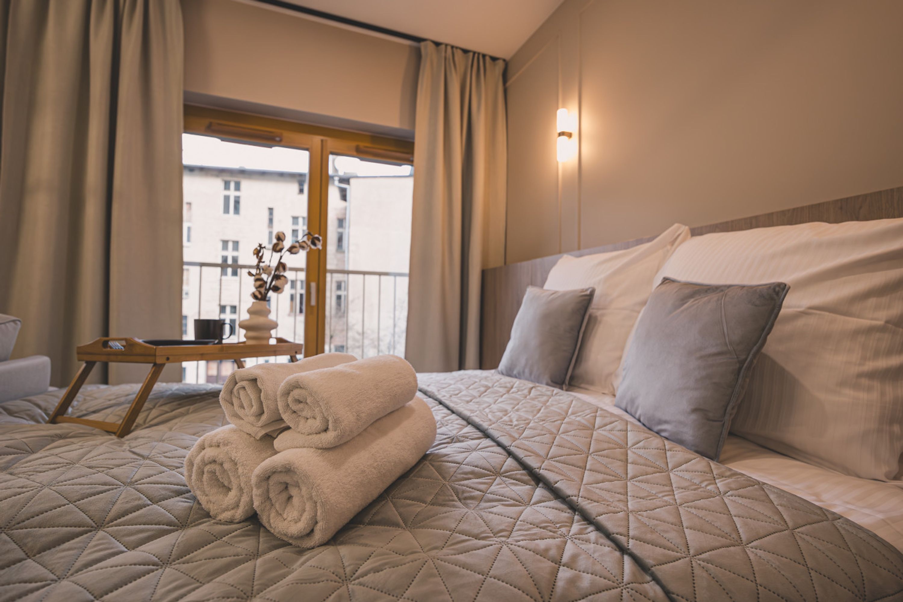 A hotel room with a large bed, a bedside table with a vase of dried flowers, and a view of buildings through the window.