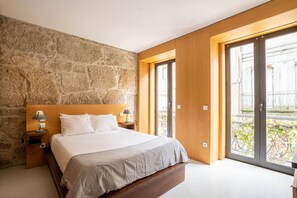 Une chambre avec un mur en pierre, une tête de lit en bois, un grand lit, deux tables de chevet avec des lampes et une porte-fenêtre coulissante donnant sur un balcon.