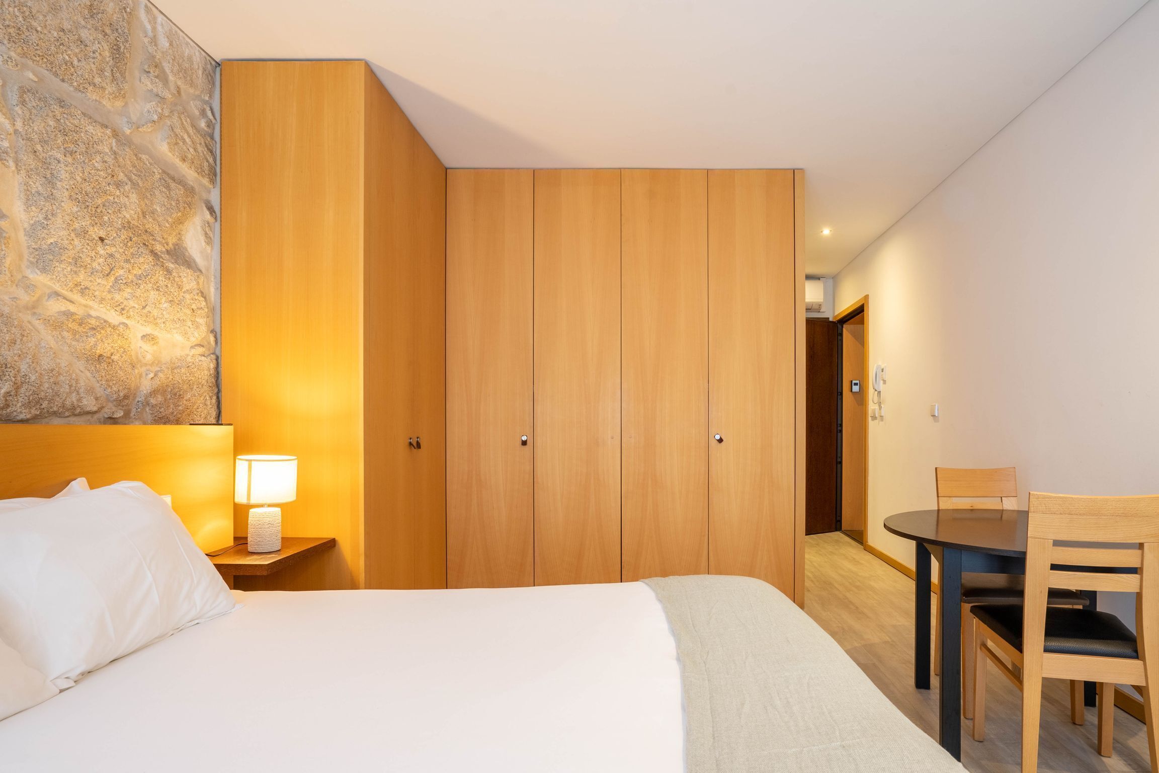 Ein Hotelzimmer mit einem Bett, einem Nachttisch mit Lampe, einem Holzschrank, einem Esstisch mit Stühlen und einem Flur, der zu einem anderen Zimmer führt.